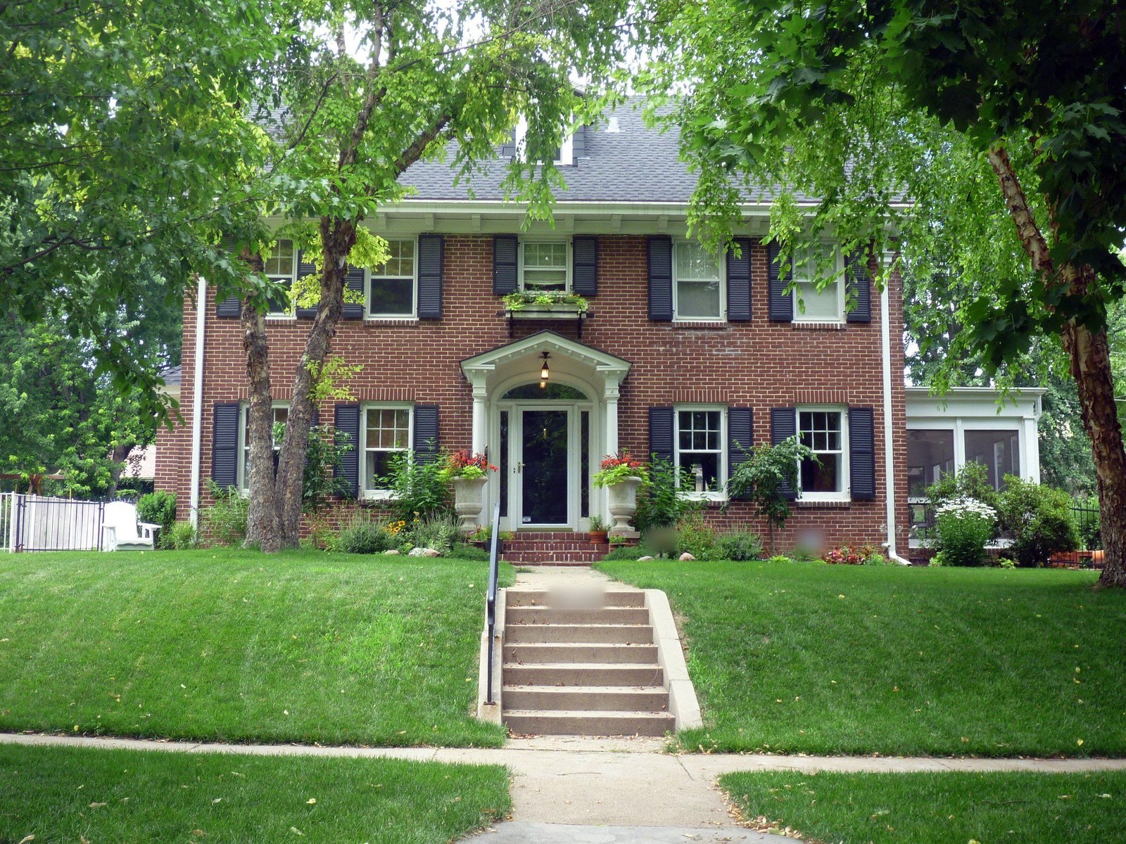 Two-story red brick colonial house with black shutters, a white front portico, and a sloped green lawn with steps up from the sidewalk, framed by trees