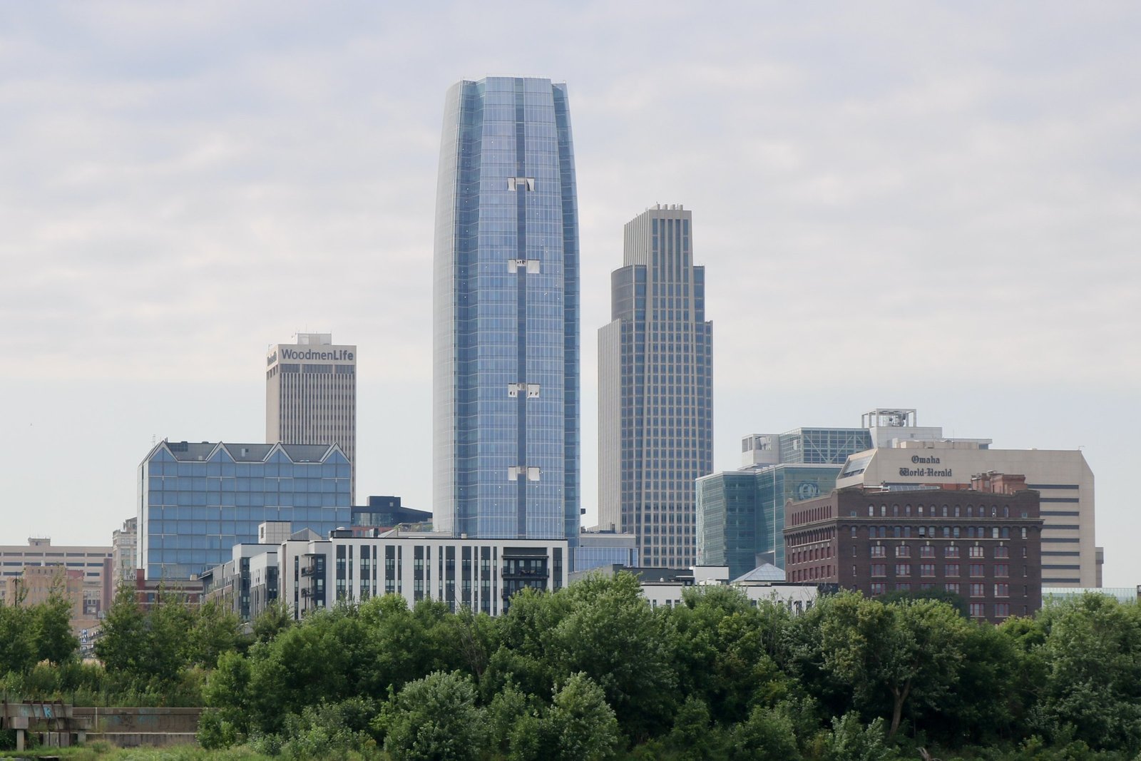 Downtown Omaha skyline under a cloudy summer sky, with a tall curved glass tower in the center, the WoodmenLife tower and the Omaha World-Herald building on either side, and a line of green trees in the foreground
