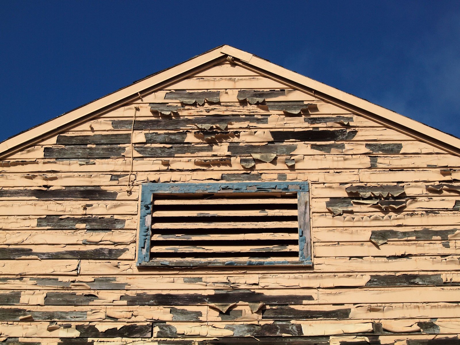 Gable of an older wooden house with peeling paint and a vent, against a clear blue sky